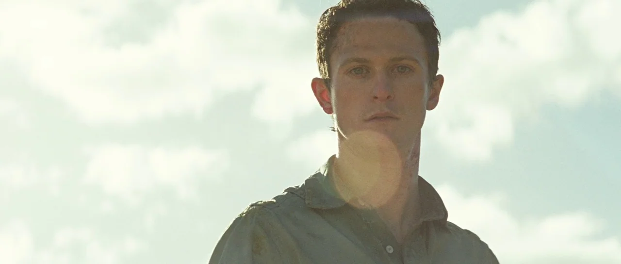 The Ruins (2008) movie still: medium — An open field under a bright sky with soft, diffused daylight. A young man stands looking forward,…