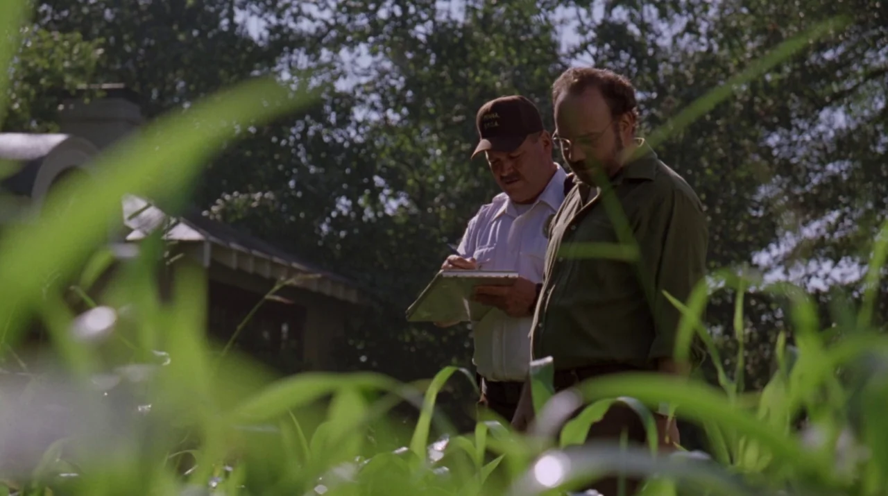 Lady in the Water (2006) movie still: medium — Two men stand in a grassy area with mature trees and a house roof visible. One man holds a clipboard…