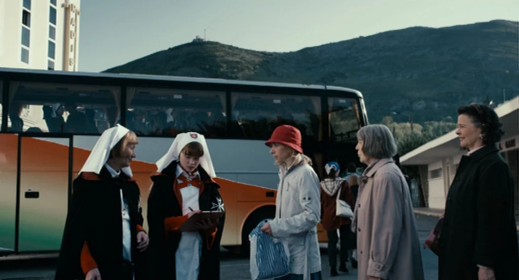 Lourdes movie still: wide — A paved lot sits beneath a mountain, featuring a large tour bus with branding. A group of women in various attire stand…