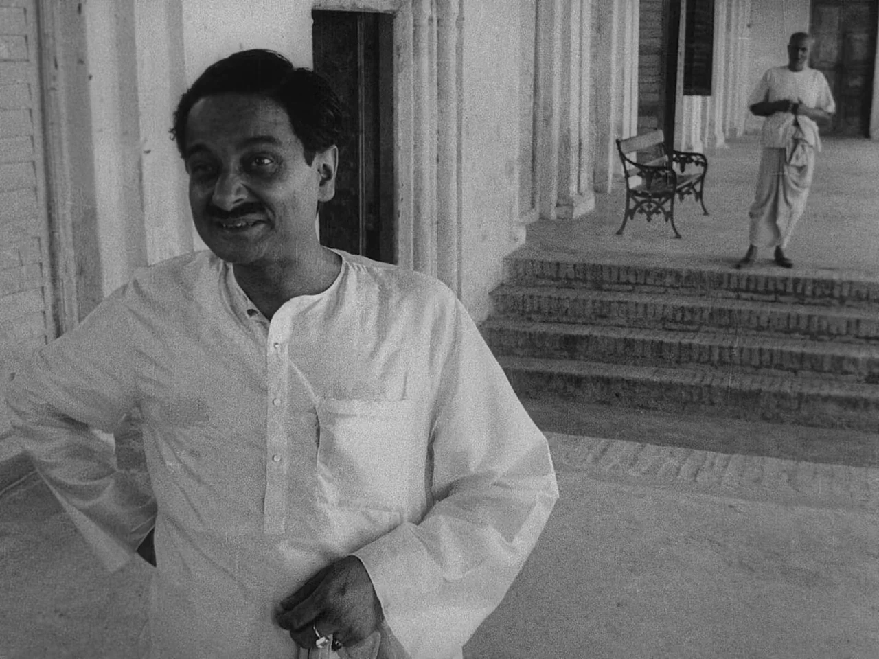 The Music Room (1958) movie still: medium — A stone veranda with fluted columns features a decorative wrought iron bench nearby. A man in a white…