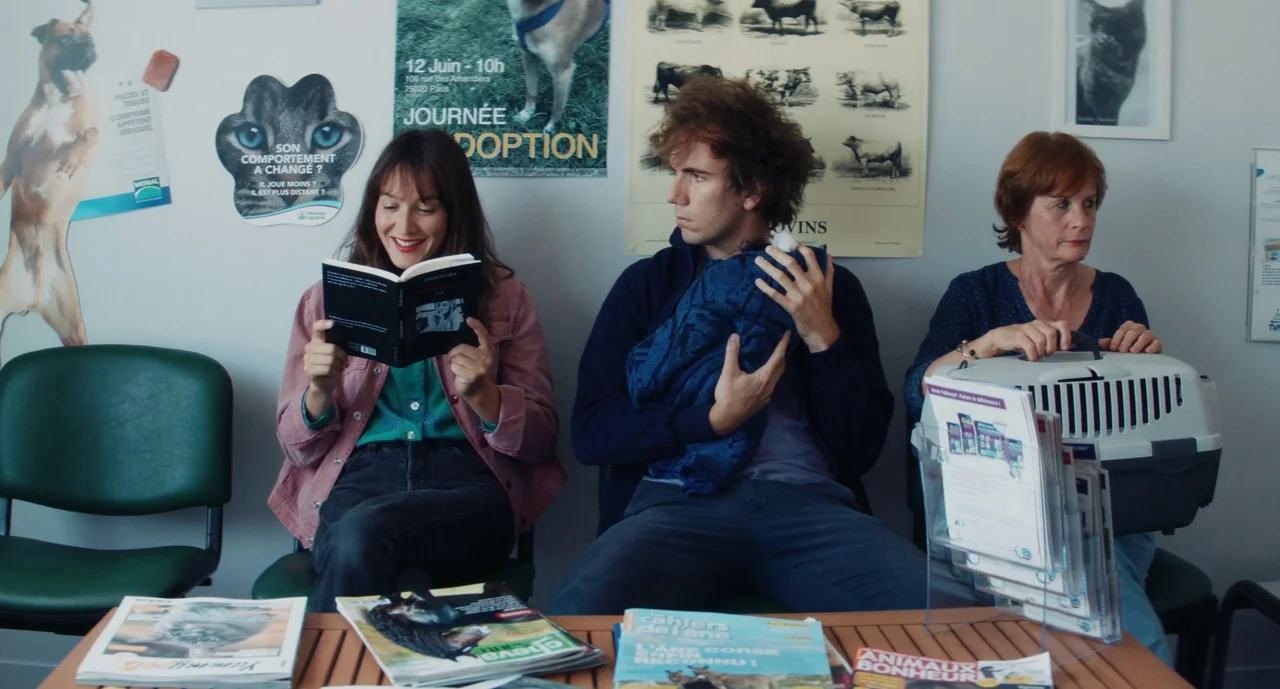 Anaïs in Love (2021) movie still: medium — A quiet veterinary clinic waiting area features posters of animals on white walls. Three people sit on…