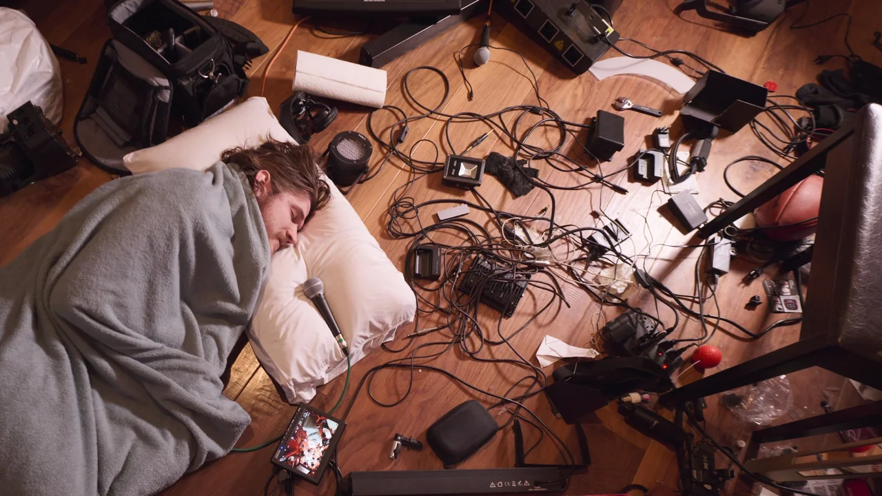 Bo Burnham: Inside (2021) movie still: wide — A wooden floor is cluttered with miscellaneous film equipment, cables, and production gear. A man lies…