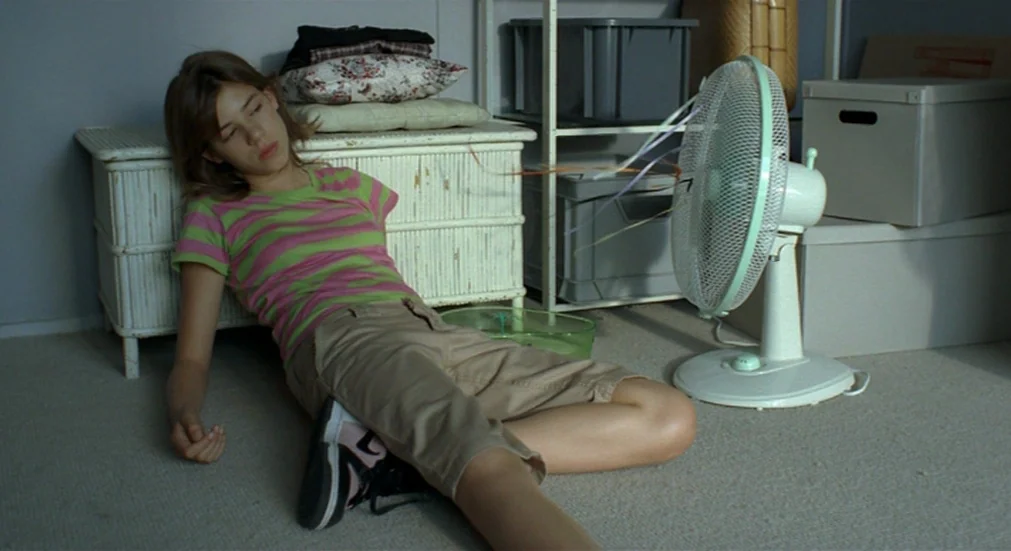 Water Lillies (2007) movie still: fullbody — A teenage girl sits on the floor of a storage room filled with boxes. She rests against a white wicker…