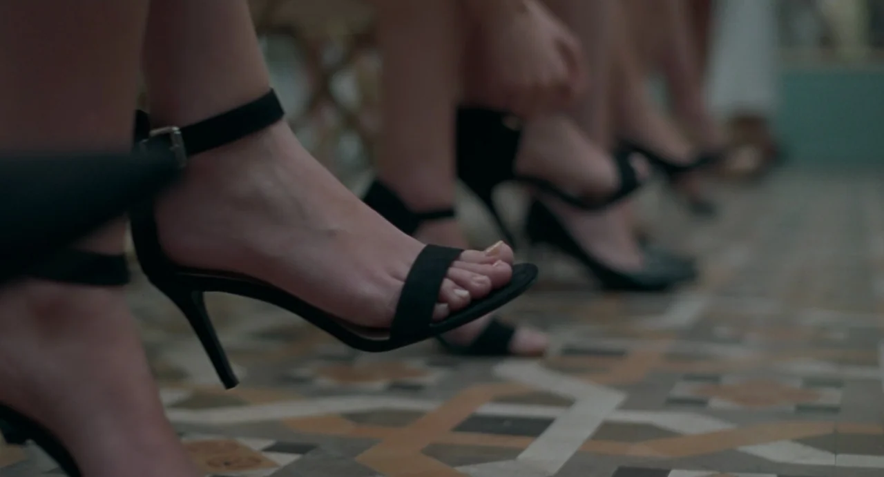 La Abuela (2021) movie still: closeup — A row of feet wearing black high-heeled sandals rests on an ornate tile floor. The background shows multiple…