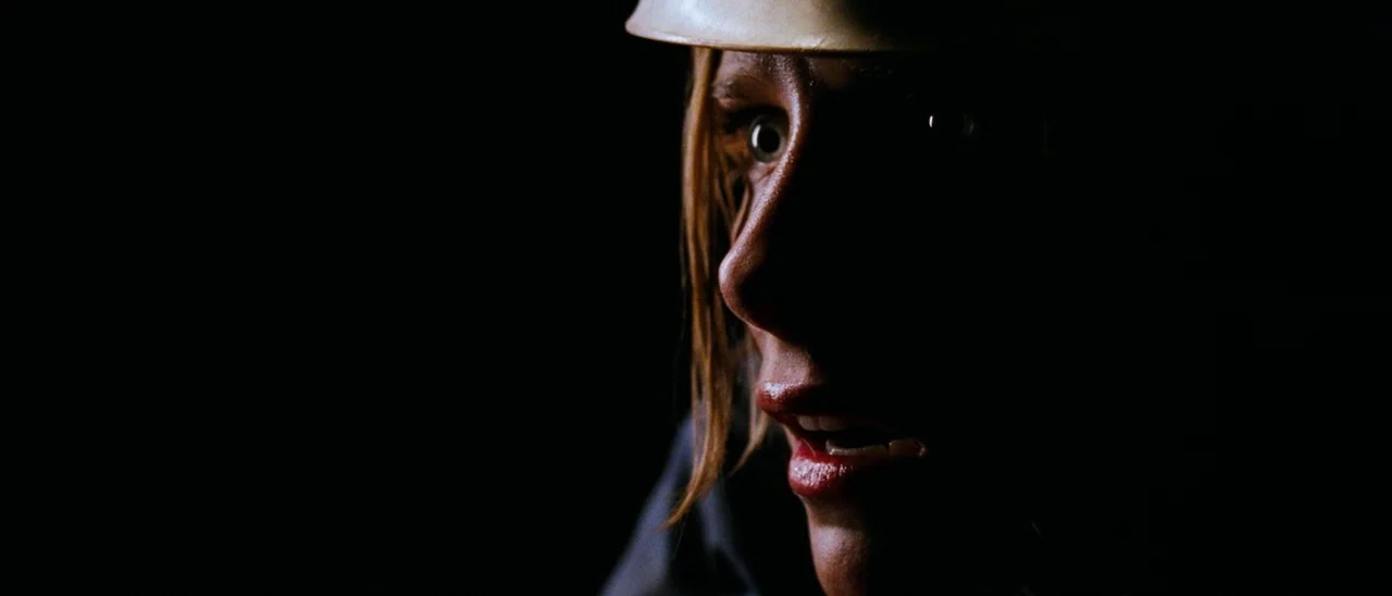 The Descent (2005) movie still: closeup — The frame shows a dark environment where the subject is illuminated by directional lighting. A woman…