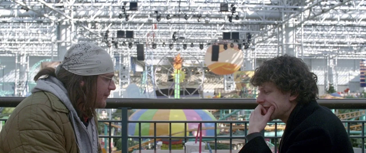 The End of the Tour (2015) movie still: medium — A food court overlooks a large indoor amusement park with white steel support structures. Two men…