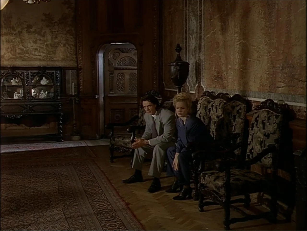 A Change of Place (1994) movie still: medium — A dark, wood-paneled room features ornate wall tapestries and antique furniture with carved details.…