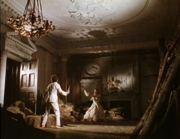 The Tempest (1979) movie still: wide — An ornate room in a grand house is filled with hay and wooden branches. A man and a woman in white attire…