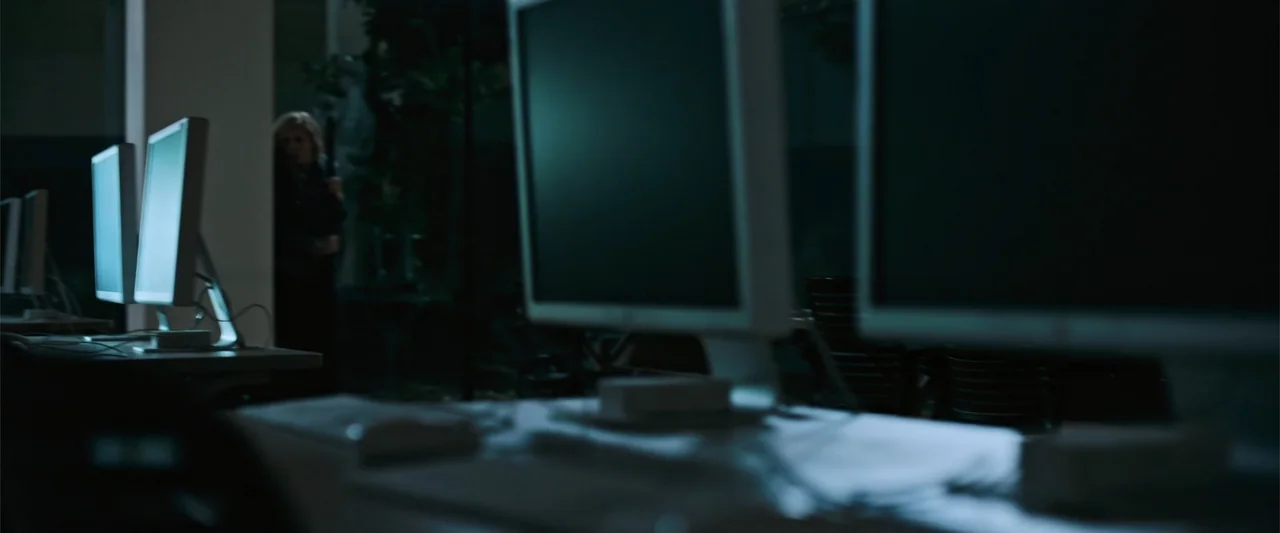 Init!ation (2021) movie still: medium — A dark, dimly lit computer lab features rows of monitors in the foreground. A lone woman stands near a…