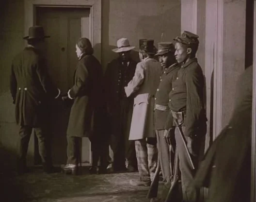 Birth of a Nation (1915) movie still: fullbody — A group of men in 19th-century attire stand in a narrow hallway. Two men in uniform guard the…