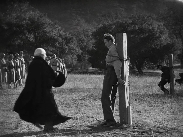 Mata Hari (1931) movie still: fullbody — An open grassy area contains wooden posts where men are bound by rope. A robed figure walks toward a…