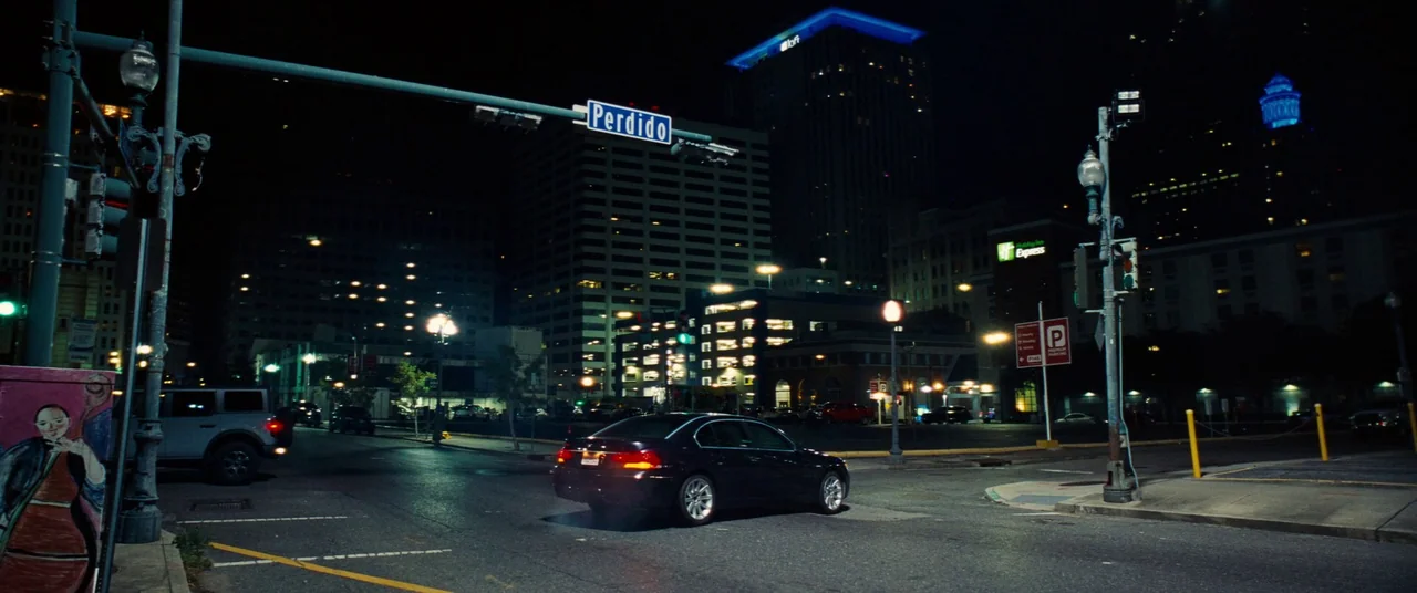 Kinds of Kindness (2024) movie still: wide — A nighttime city street intersection features a sedan driving past a Perdido street sign. Tall…