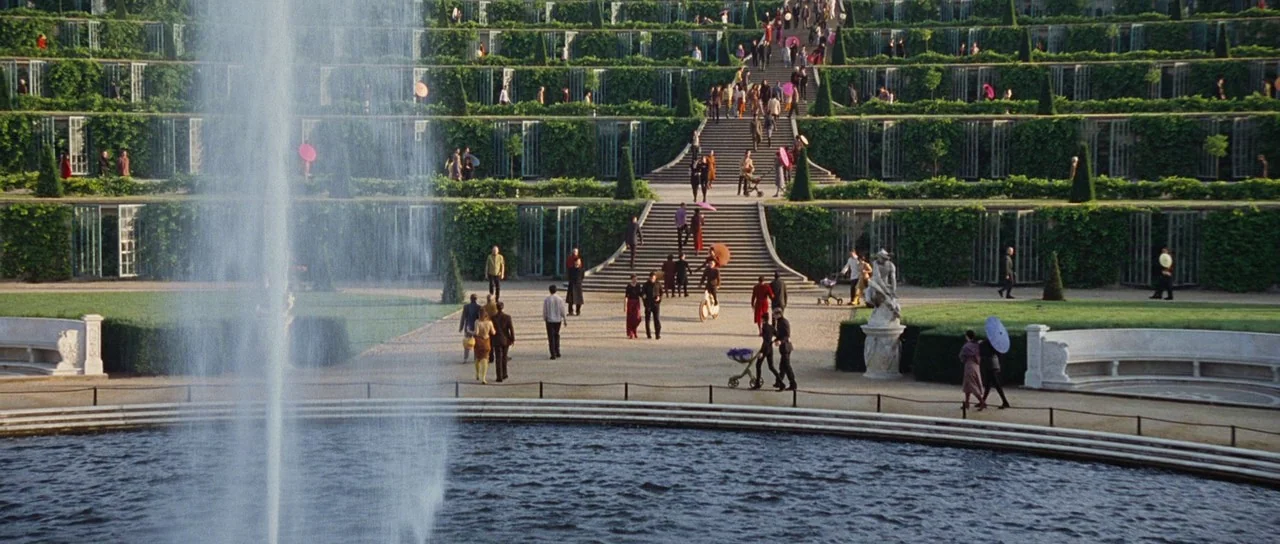 Aeon Flux (2005) movie still: establishing — A expansive formal park features tiered stone staircases lined with green hedges and trees. Many people…