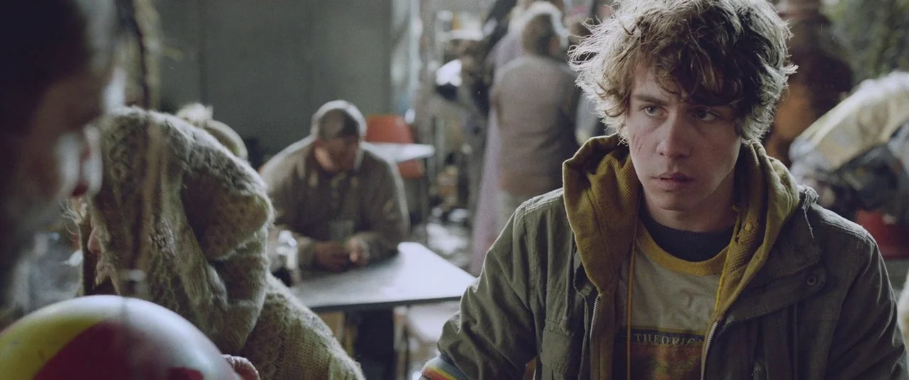 Turbo Kid (2015) movie still: medium — A cluttered communal space with various individuals sitting at tables in the blurred background. A young man…