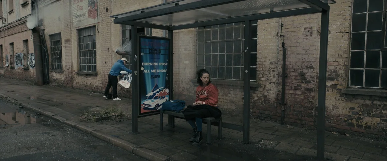 Perfect Addiction (2023) movie still: wide — A weathered brick wall with industrial windows sits behind a metal and glass bus shelter. A young woman…