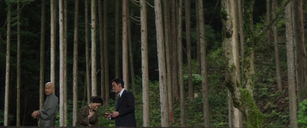 The Handmaiden (2016) movie still: wide — A dense grove of tall, thin cedar trees with moss-covered ground and tree trunks. Three people in formal…
