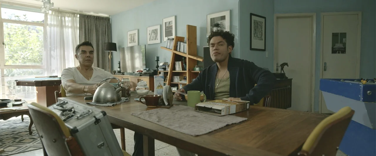 Divine Intervention (2026) movie still: medium — A well-lit contemporary residential room with light blue walls and wooden furniture. Two men sit at…