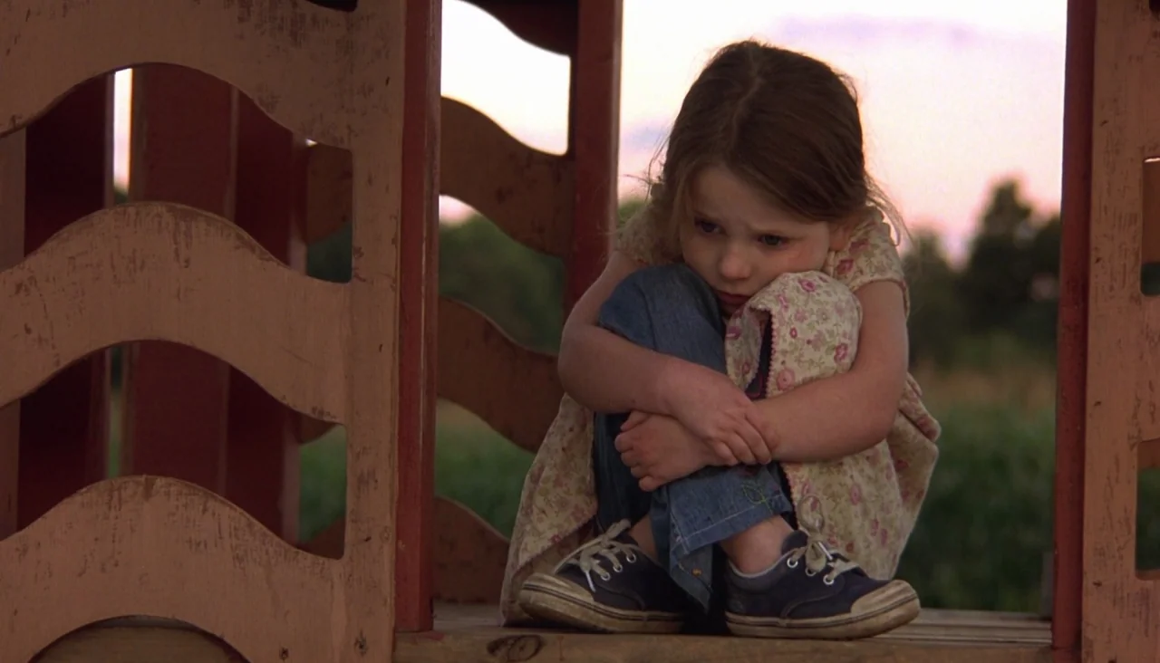 Signs (2002) movie still: medium — A wooden structure provides a small shelter for a child in an outdoor setting. The young girl sits alone with her…