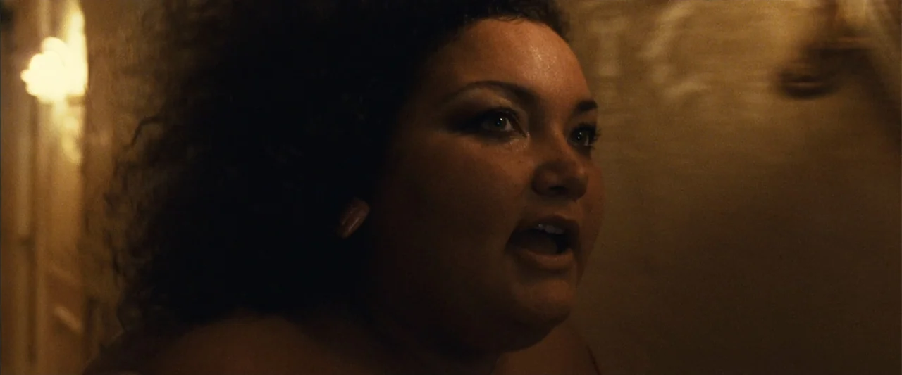 Reality (2023) movie still: closeup — A dimly lit indoor hallway with warm artificial lighting on a textured wall. A woman with dark curly hair…