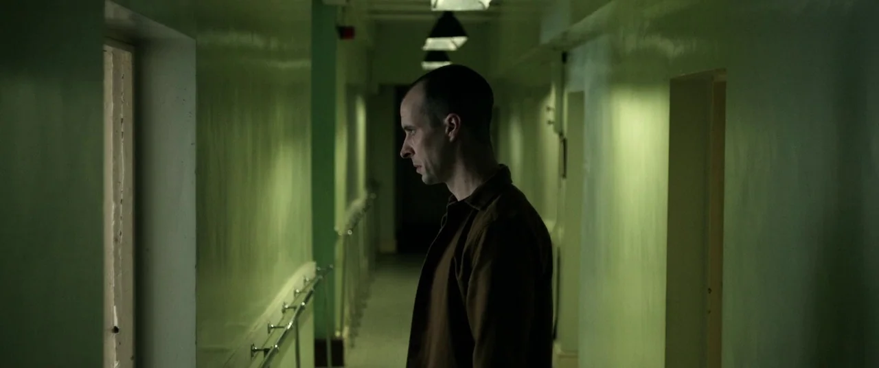 The Cured (2017) movie still: medium — A long, narrow corridor features mint-green walls and a series of identical closed doors. A man in a brown…