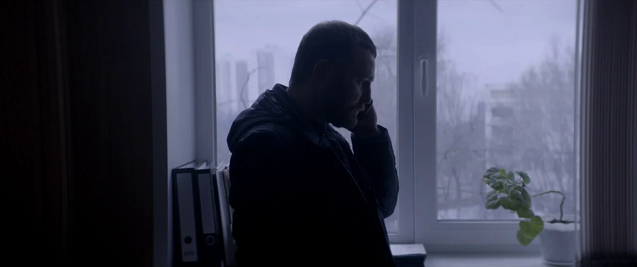 Loveless (2017) movie still: medium — A man stands before a large window looking out at a bleak urban landscape. He is talking on a mobile phone in…