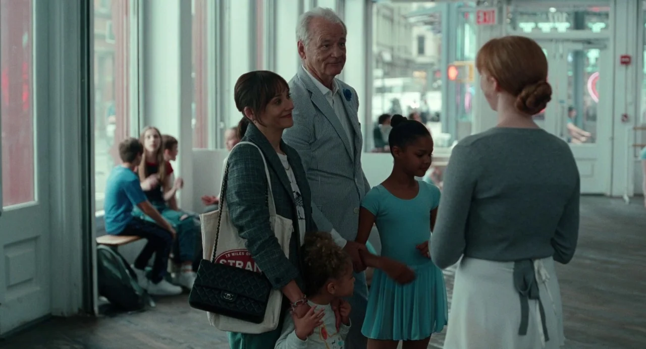 On The Rocks (2020) movie still: medium — An airy room with large street-facing windows showing a busy city exterior. A group of adults and children…
