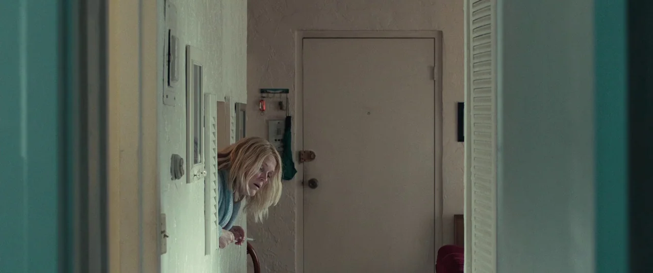 Stronger (2017) movie still: medium — A muted beige indoor wall features a rectangular cutout window between two rooms. A blonde woman leans through…