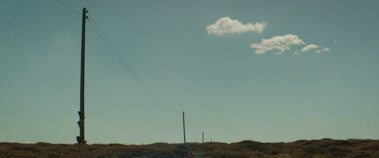Beast (2017) movie still: establishing — A flat, grassy field stretches toward the horizon under a clear, pale blue sky. Three utility poles stand…