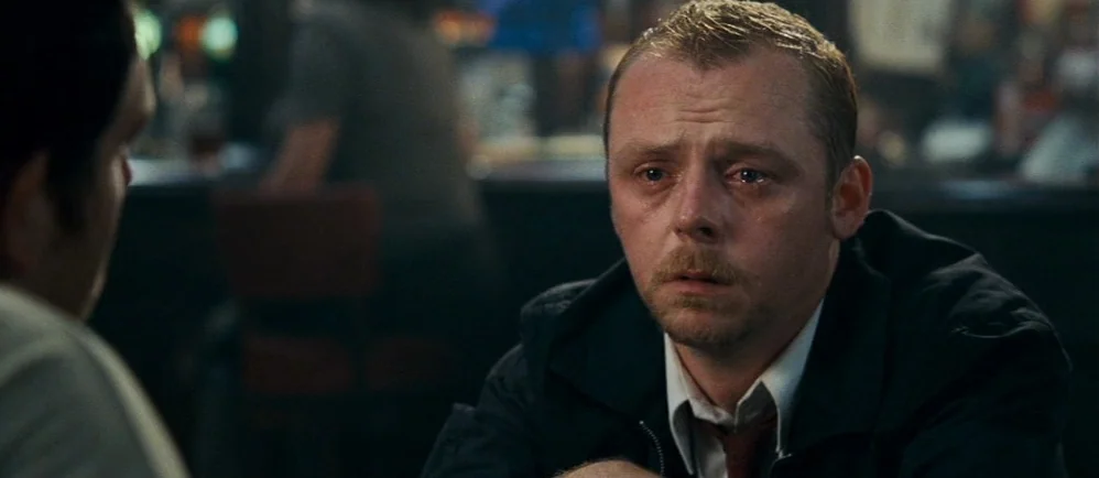 Shaun Of The Dead (2004) movie still: medium — A dimly lit indoor space with blurred background activity suggesting a late evening setting. A man…