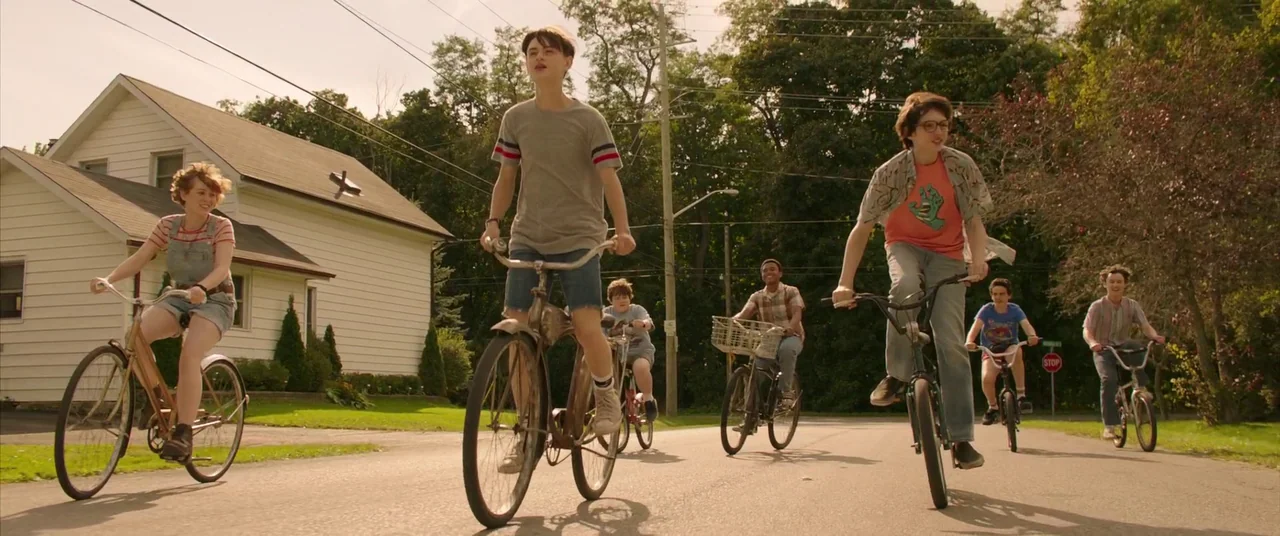 It Chapter Two (2019) movie still: wide — A group of teenagers ride bicycles down a sunlit suburban street past a white house. They move in a…