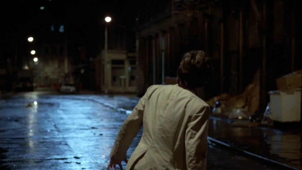 After Hours (1985) movie still: medium — A wet, dark city street at night illuminated by distant overhead lamps. A person in a beige jacket stands…