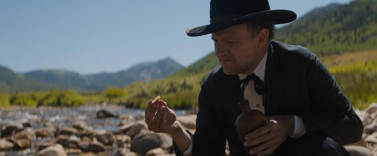Damsel (2018) movie still: medium — A rocky riverbank is bordered by lush green hills and distant mountain peaks. A man in period western clothing…