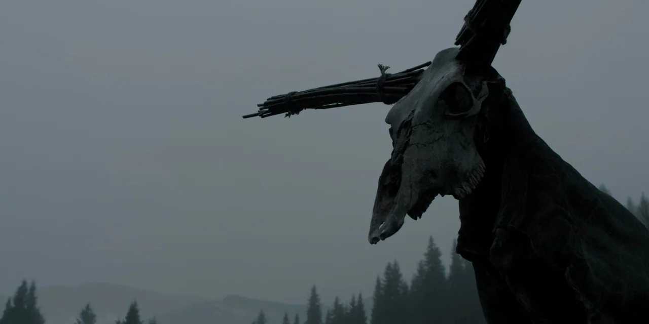 The Ritual (2017) movie still: medium — A dark, weathered animal skull is tied to a wooden stake against a sky. A distant, blurred evergreen forest…