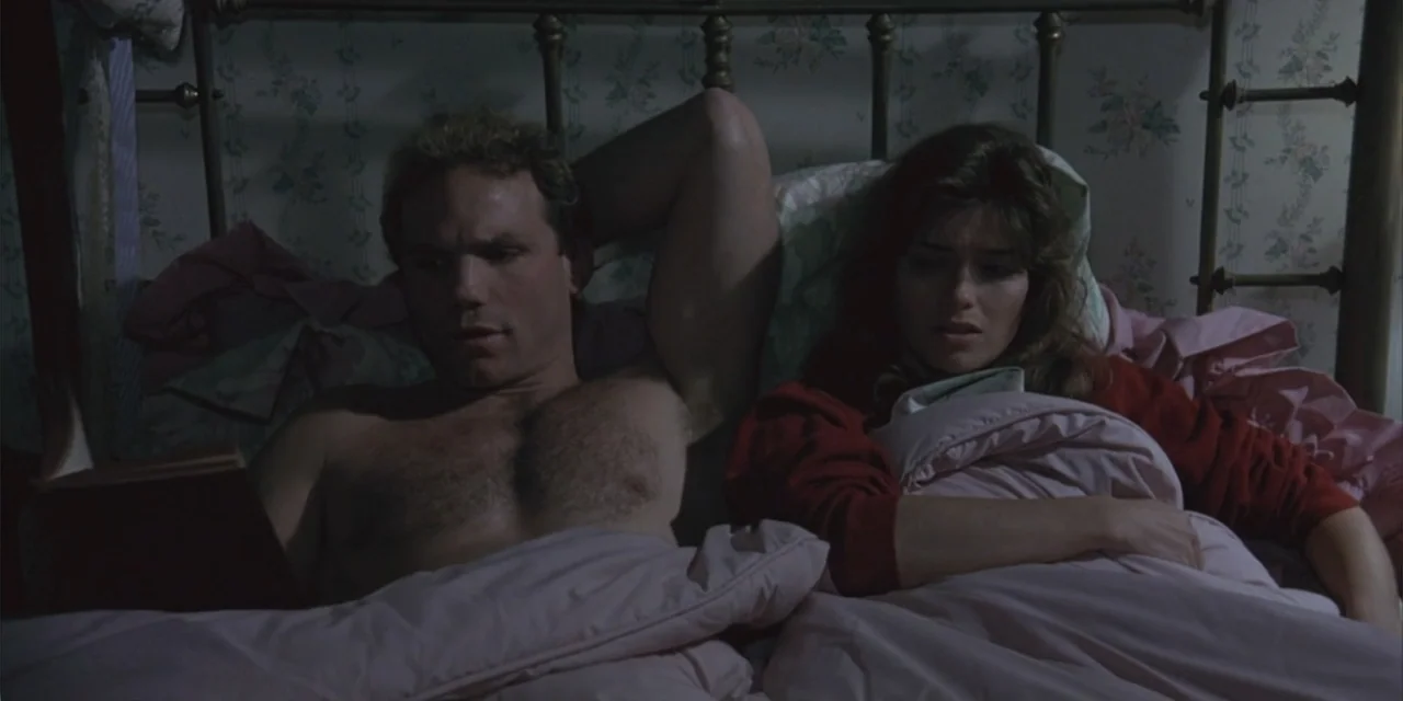 El sótano (2024) movie still: medium — A dark bedroom features a vintage iron bed frame against floral patterned wallpaper. A man reclines in bed…