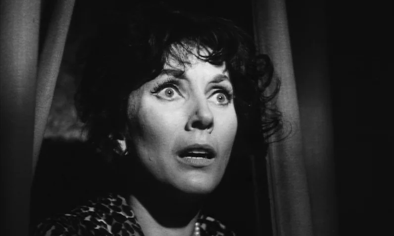 The Girl Who Knew Too Much (1963) movie still: closeup — A dark indoor setting framed by heavy curtains on either side. A woman with dark hair looks…