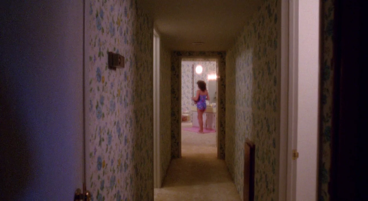 White Of The Eye (1987) movie still: wide — A long, dark hallway is covered in light blue floral wallpaper and beige carpeting. A woman stands in a…