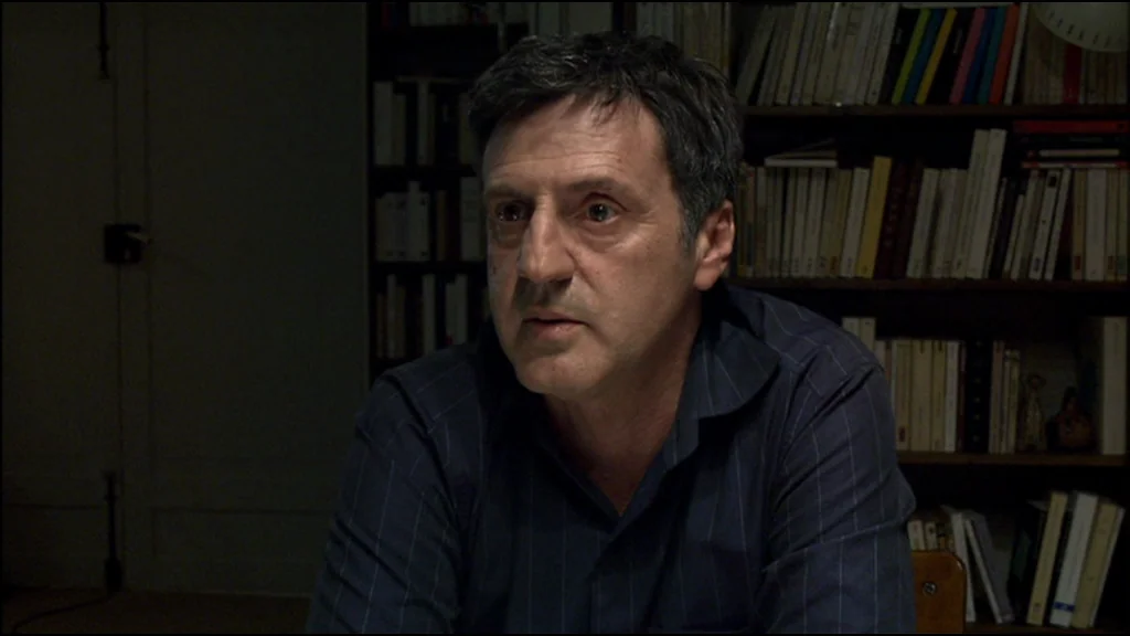 Caché (Hidden) (2005) movie still: medium — A dimly lit home office space is lined with rows of wooden bookshelves. A middle-aged man in a dark…