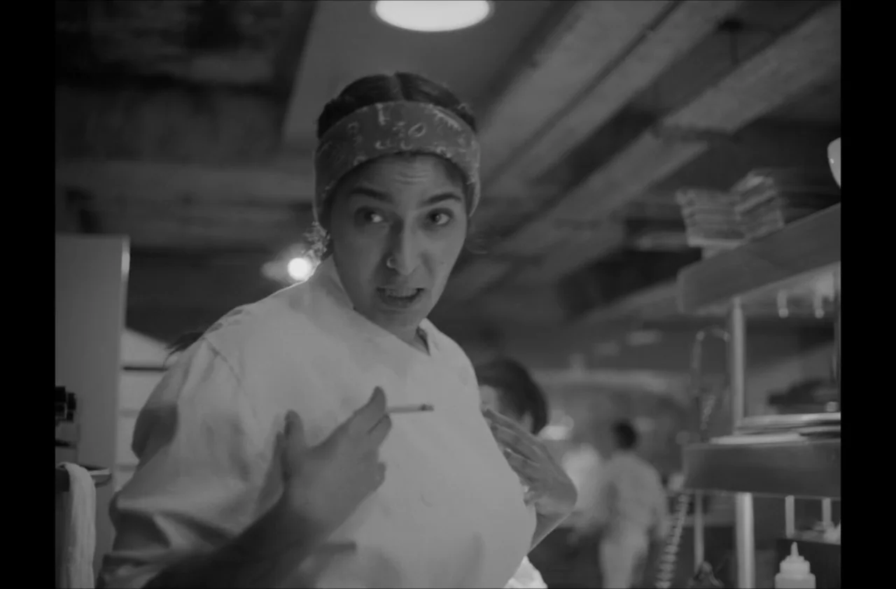The Kitchen (2012) movie still: medium — A busy commercial restaurant kitchen features stainless steel surfaces and low ceiling rafters. A female…