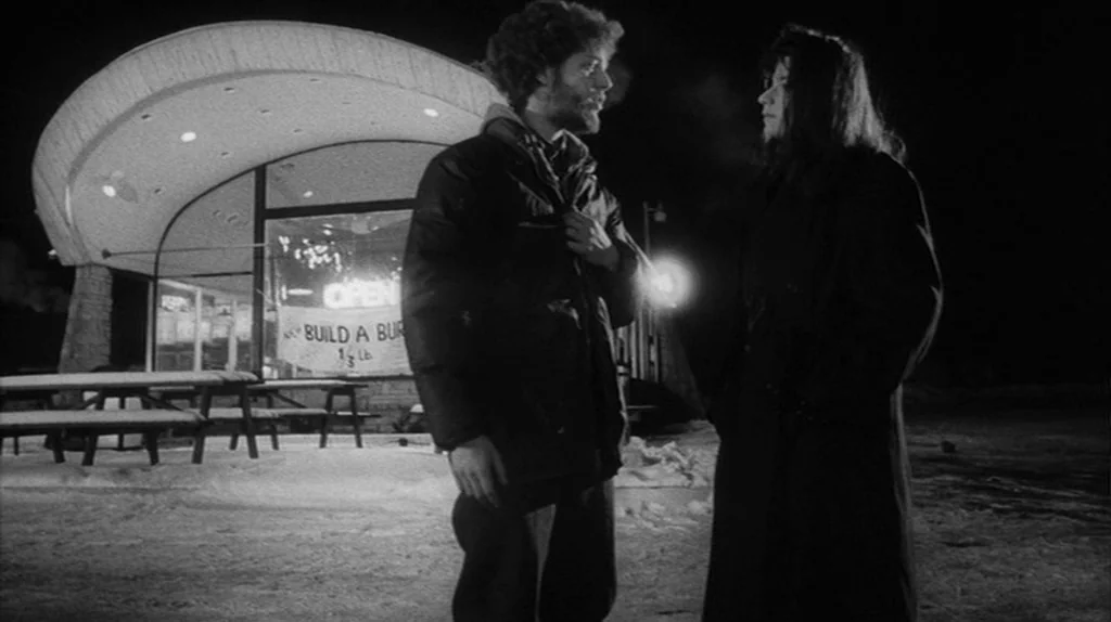 Stranger Than Paradise (1984) movie still: medium — A snowy parking lot at night sits in front of a brightly lit diner. A man and a woman stand near…