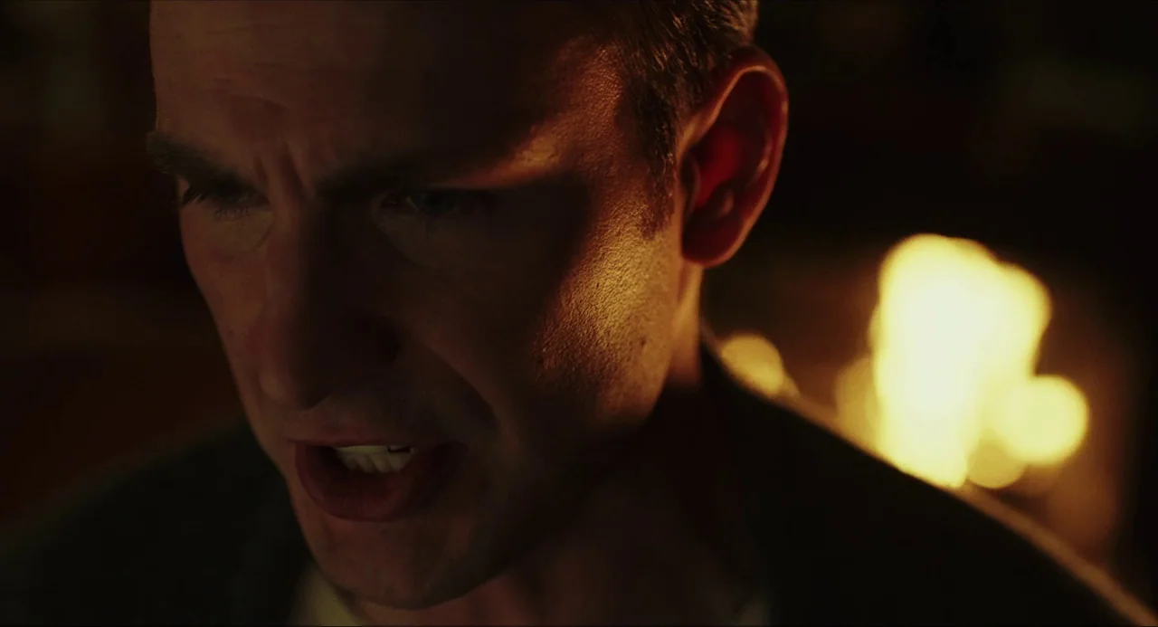 Knives Out (2019) movie still: closeup — A dark indoor space is lit by a nearby flickering fire. A man stares forward with a focused and tense…