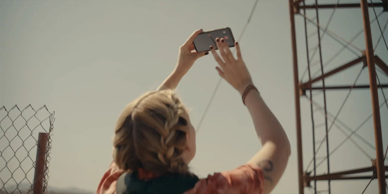 Fall (2022) movie still: medium — An outdoor area with a chain-link fence and a large metal transmission tower. A young woman with braided hair…