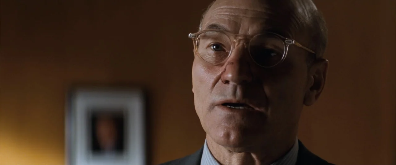 Conspiracy Theory (1997) movie still: closeup — A dimly lit office features an out-of-focus framed picture on the wall behind. An elderly bald man…