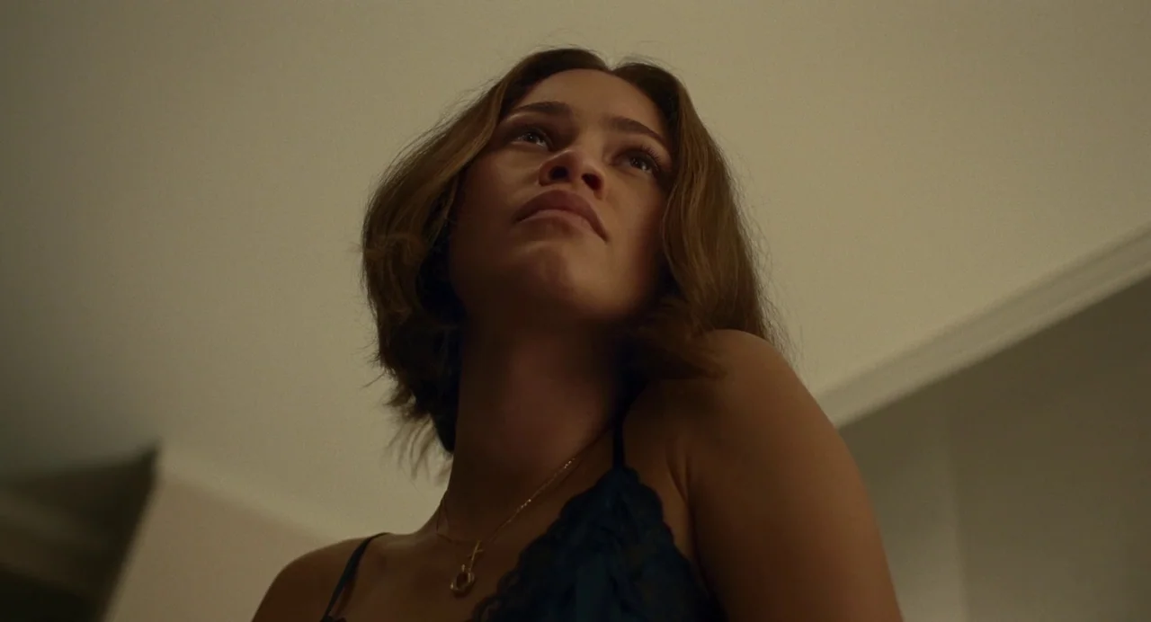 Challengers (2024) movie still: closeup — A dimly lit indoor space with plain walls and soft ceiling lighting. A woman gazes upward toward the…