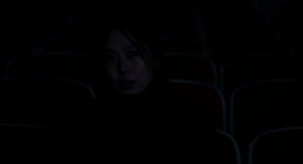 On The Beach At Night Alone (2017) movie still: medium — A dark movie theater with rows of empty, dimly lit red seats behind the subject. A woman…