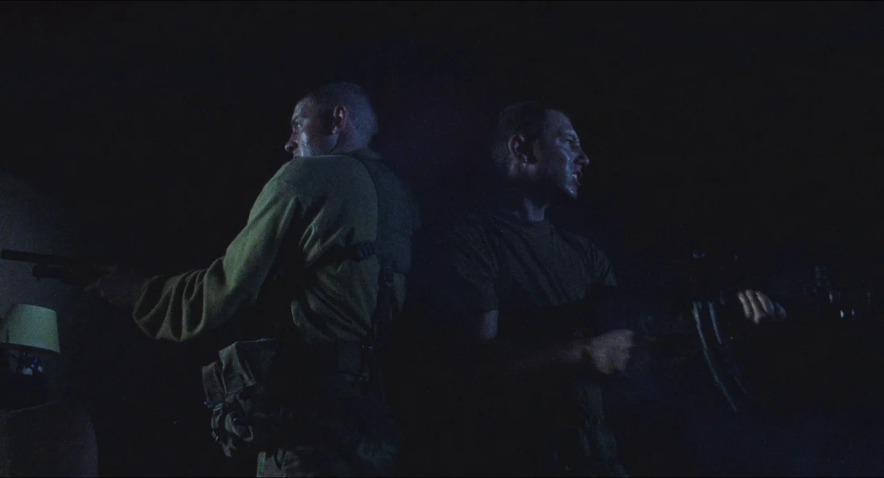 Dog Soldiers (2002) movie still: medium — Two men stand back to back in a dark, sparse room at night. They are armed with rifles and maintain a…