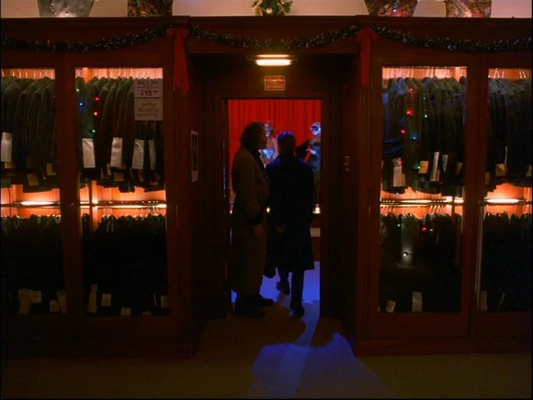 Eyes Wide Shut (1999) movie still: wide — A dimly lit clothing shop interior features wooden cabinets filled with hanging garments. Two men stand in…