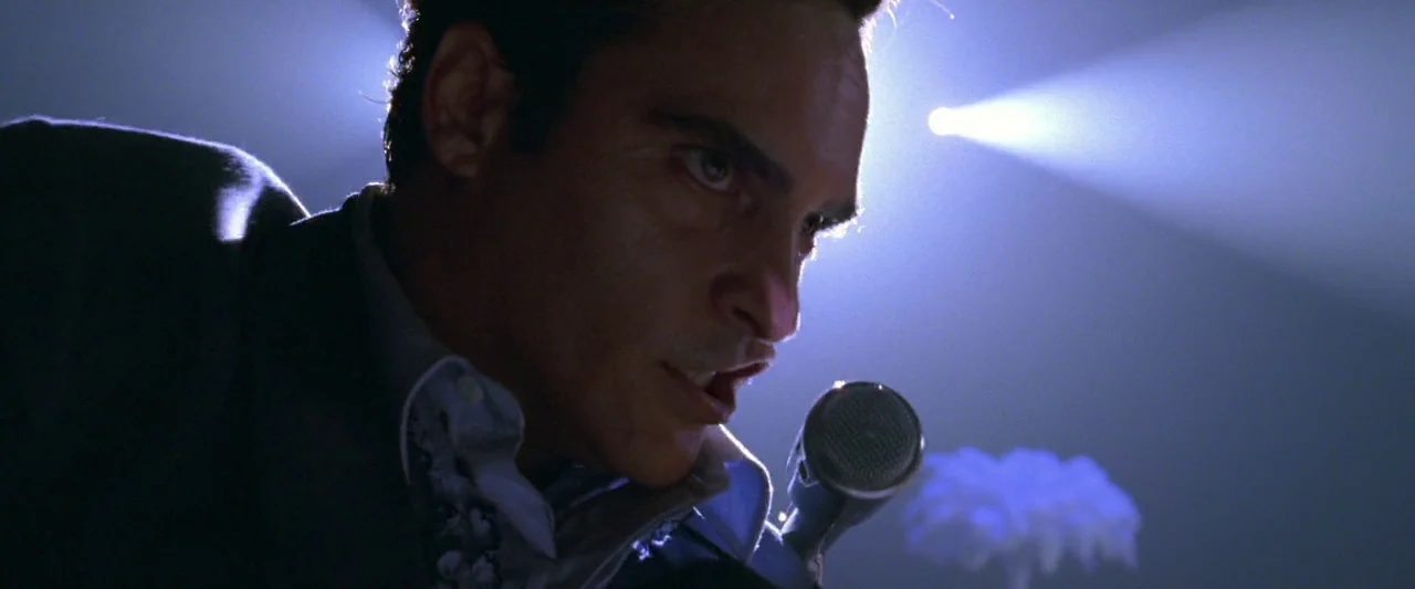 Walk The Line (2005) movie still: closeup — A dark, moody nightclub stage is illuminated by a singular, harsh focused beam. A man in a suit sings…