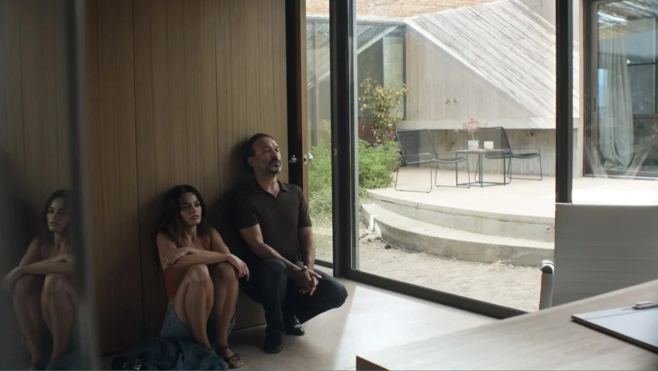 Hot Milk (2025) movie still: medium — A modern interior space features wood paneling and a large glass wall opening outward. A man and a woman sit…