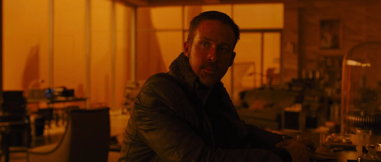 Blade Runner 2049 (2017) movie still: medium — A dark, dimly furnished interior space with floor-to-ceiling windows showing an orange exterior haze.…