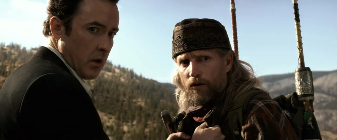 2012 (2009) movie still: medium — A mountainous outdoor landscape with blurred evergreen trees visible in the background. Two men stand close…